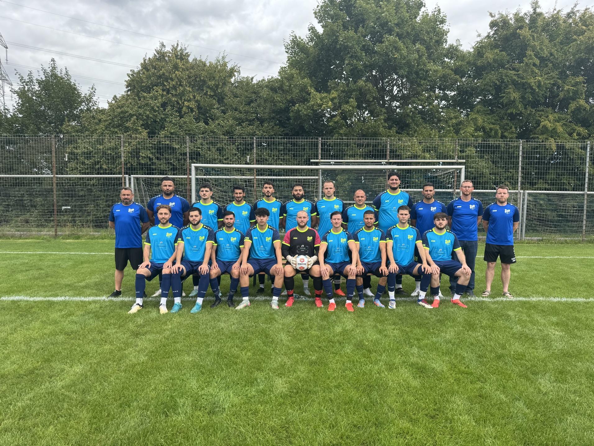 Mannschaftsfoto einer Fu&szlig;ballmannschaft mit 20 Spielern und Trainern auf einem Rasenplatz vor einem Tor.
