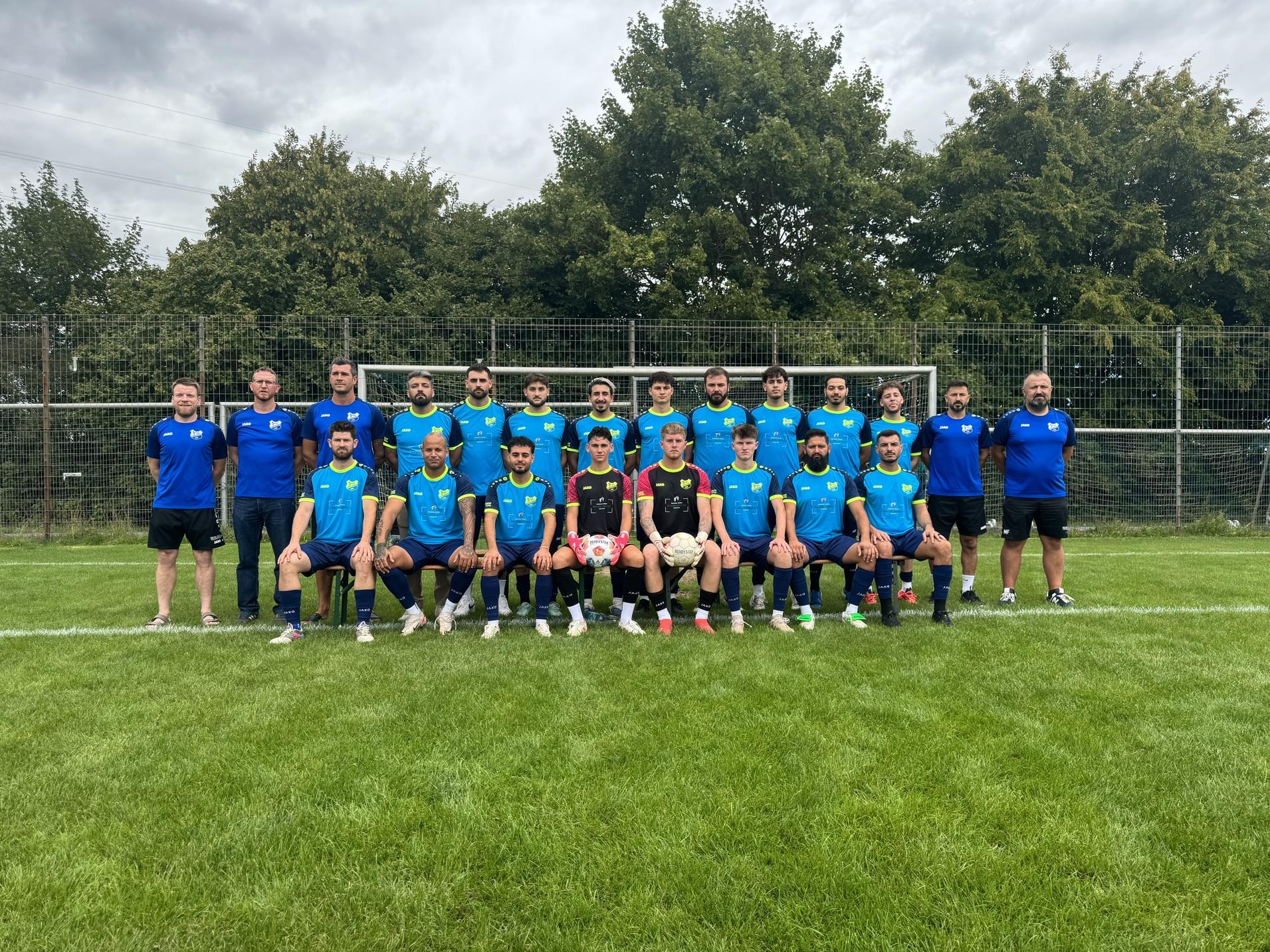Mannschaftsfoto einer Fu&szlig;balltruppe in blauen Trikots auf einem Rasenplatz mit Toren im Hintergrund.