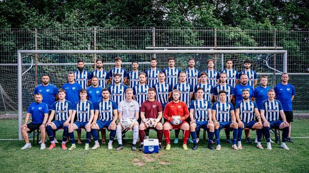 Mannschaftsaufstellung mit 28 Fu&szlig;ballspielern in blauen und gestreiften Trikots vor einem Tornetz auf einem Rasenfeld.
