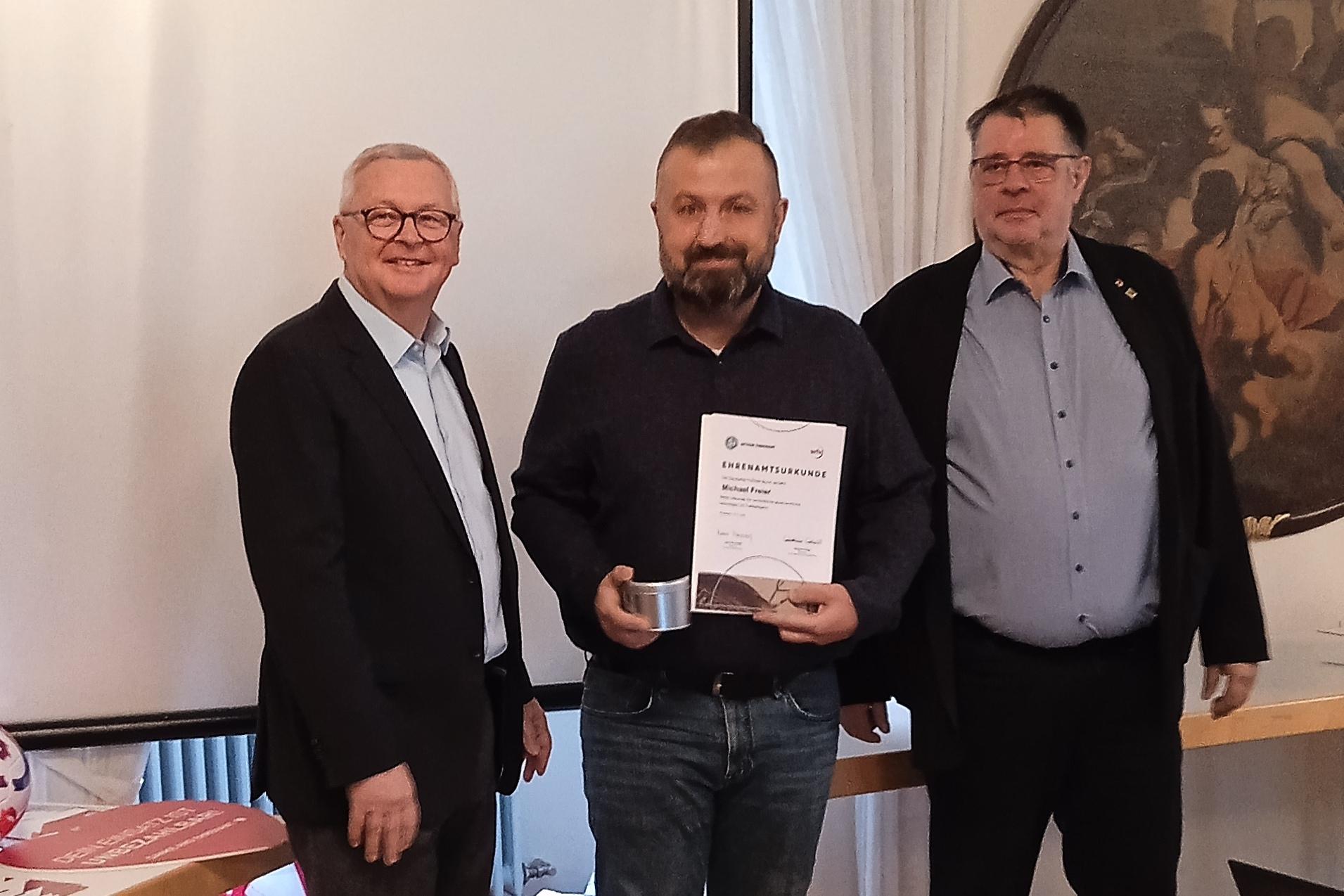 Drei M&auml;nner stehen in einem Raum, einer h&auml;lt ein Zertifikat, w&auml;hrend die anderen l&auml;cheln und zuschauen.
