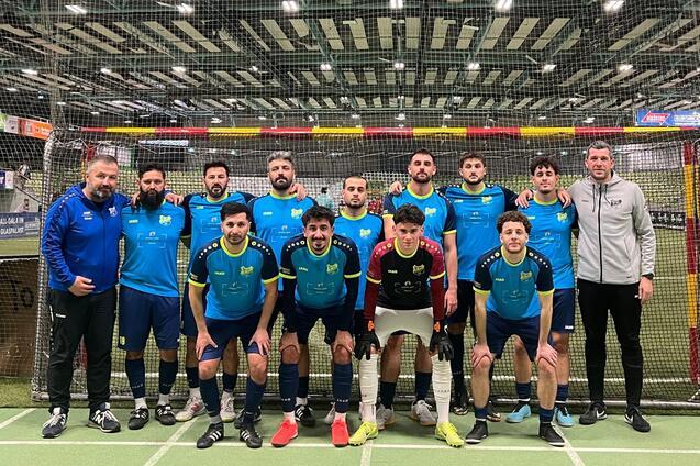 Gruppenzusammenstellung von zw&ouml;lf Fu&szlig;ballspielern in blauen Trikots vor einem Tor in einer Sporthalle.