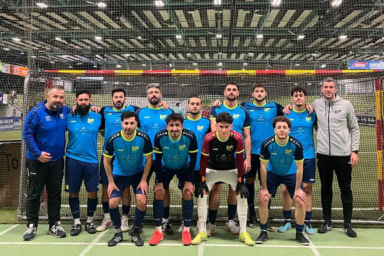 Gruppenzusammenstellung von zw&ouml;lf Fu&szlig;ballspielern in blauen Trikots vor einem Tor in einer Sporthalle.