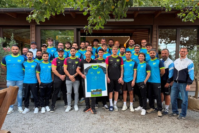 Gruppenfoto einer Sportmannschaft in blauen Trikots vor einem Holzgeb&auml;ude umgeben von B&auml;umen.