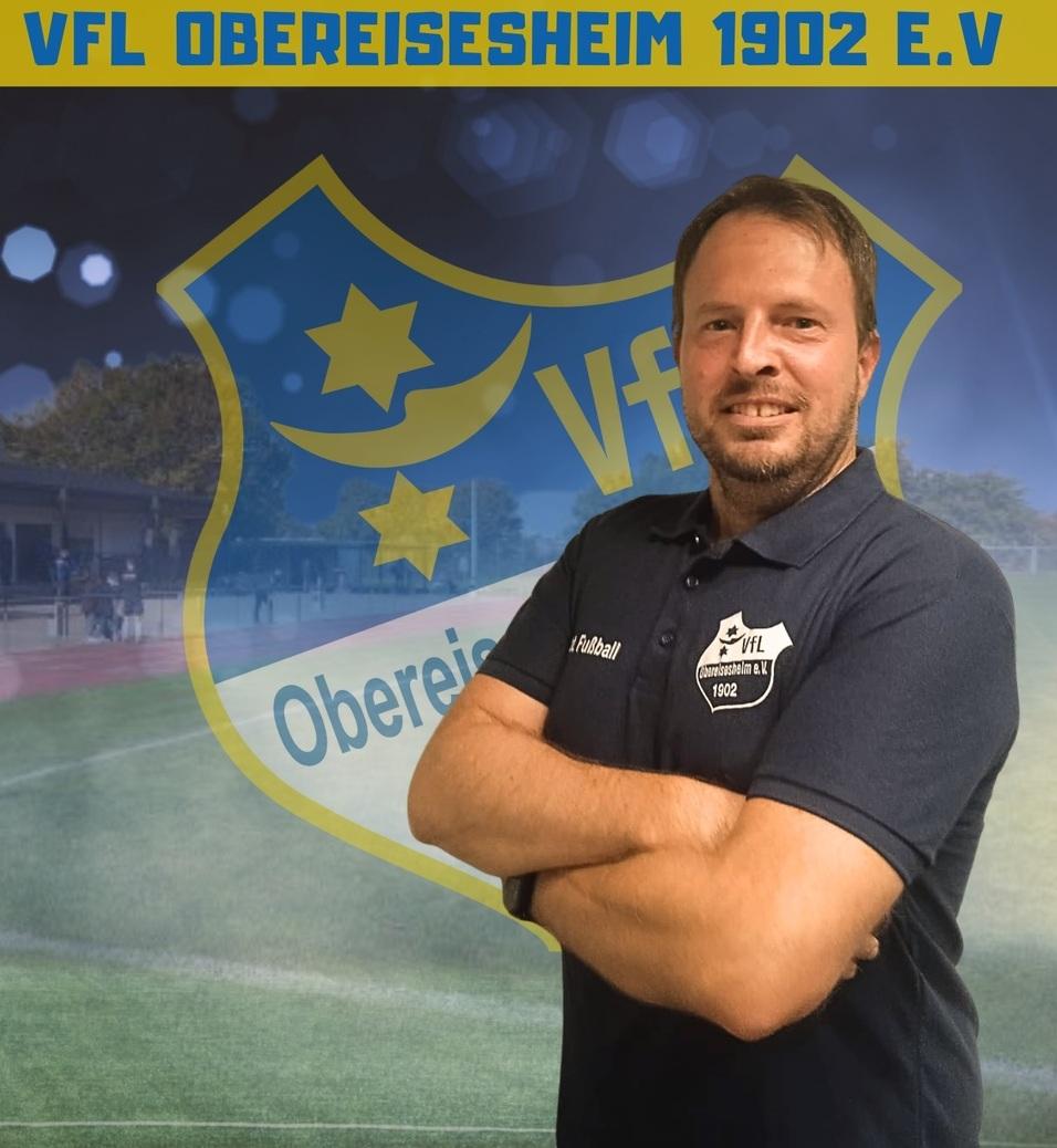 Mann in Sportbekleidung steht vor einem Vereinslogo des VfL Obereisesheim 1902 e.V. mit Fu&szlig;ballfeld im Hintergrund.