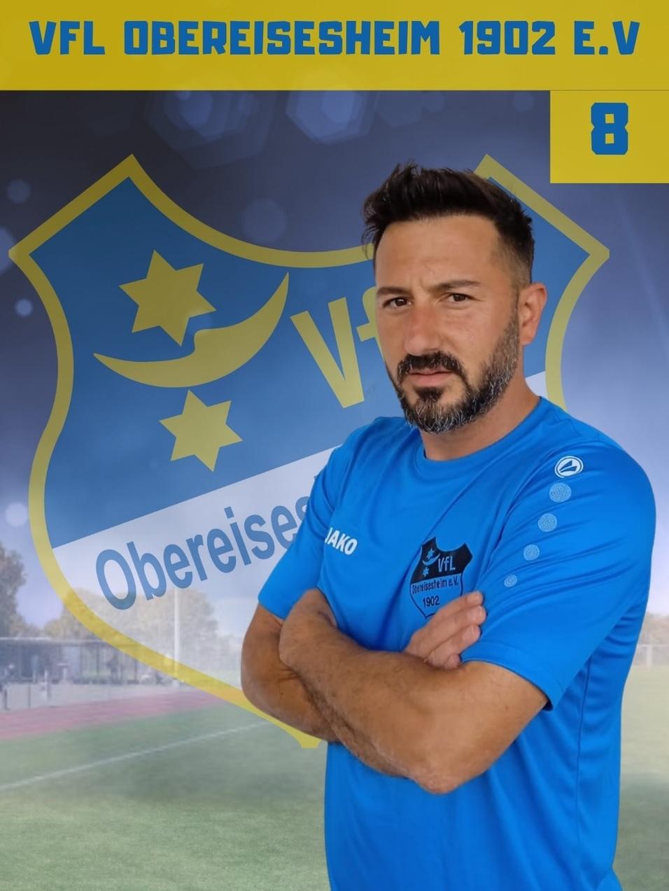 Portr&auml;t eines Mannes in blauem Sportshirt mit verschr&auml;nkten Armen vor einem Vereinswappen des VFL Obereisesheim 1902.