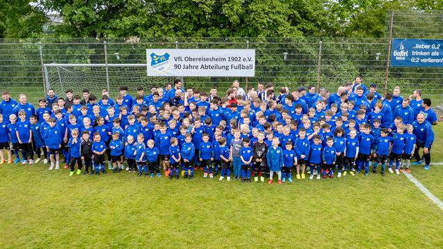 Gruppenfoto von Kindern und Jugendlichen in blauen Trikots auf einem Fu&szlig;ballplatz, umgeben von B&auml;umen und einem Banner.