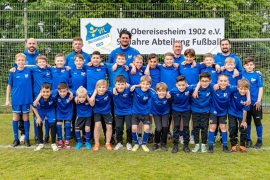 Gruppenfoto von jungen Fu&szlig;ballspielern in blauen Trikots und Trainern vor einem Vereinsbanner auf dem Fu&szlig;ballplatz.