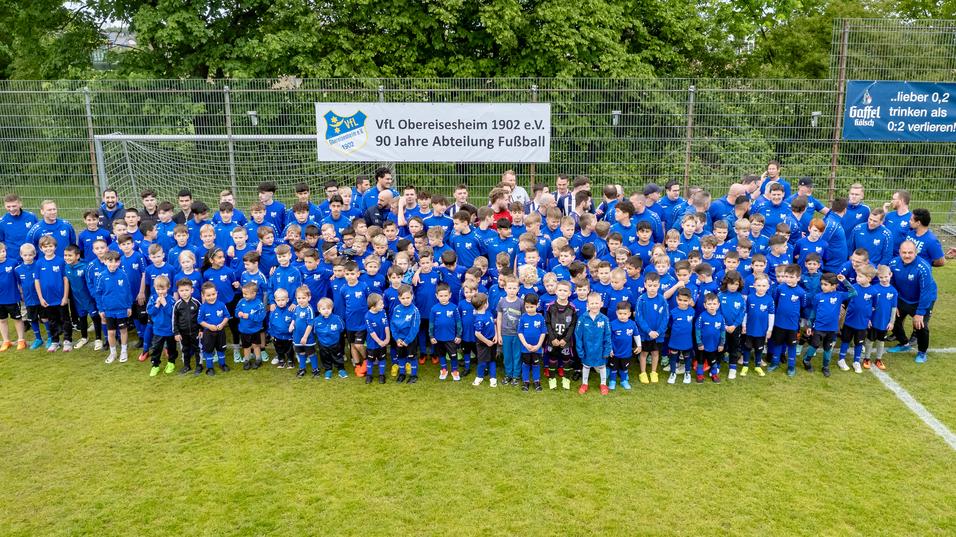 Gruppenfoto von Kindern und Jugendlichen in blauen Trikots auf einem Fu&szlig;ballplatz, umgeben von B&auml;umen und einem Banner.