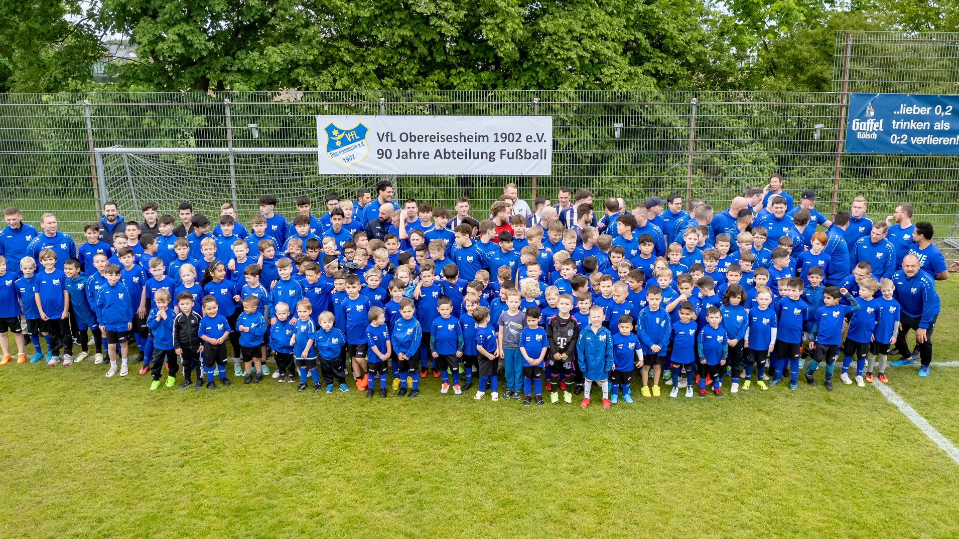 Gruppenfoto von Kindern und Jugendlichen in blauen Trikots auf einem Fu&szlig;ballplatz, umgeben von B&auml;umen und einem Banner.