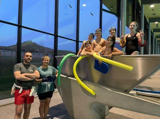 Gruppe von sechs Kindern in Schwimmkleidung und zwei Erwachsenen am Rand eines Schwimmbeckens mit Wasser-Nudeln.