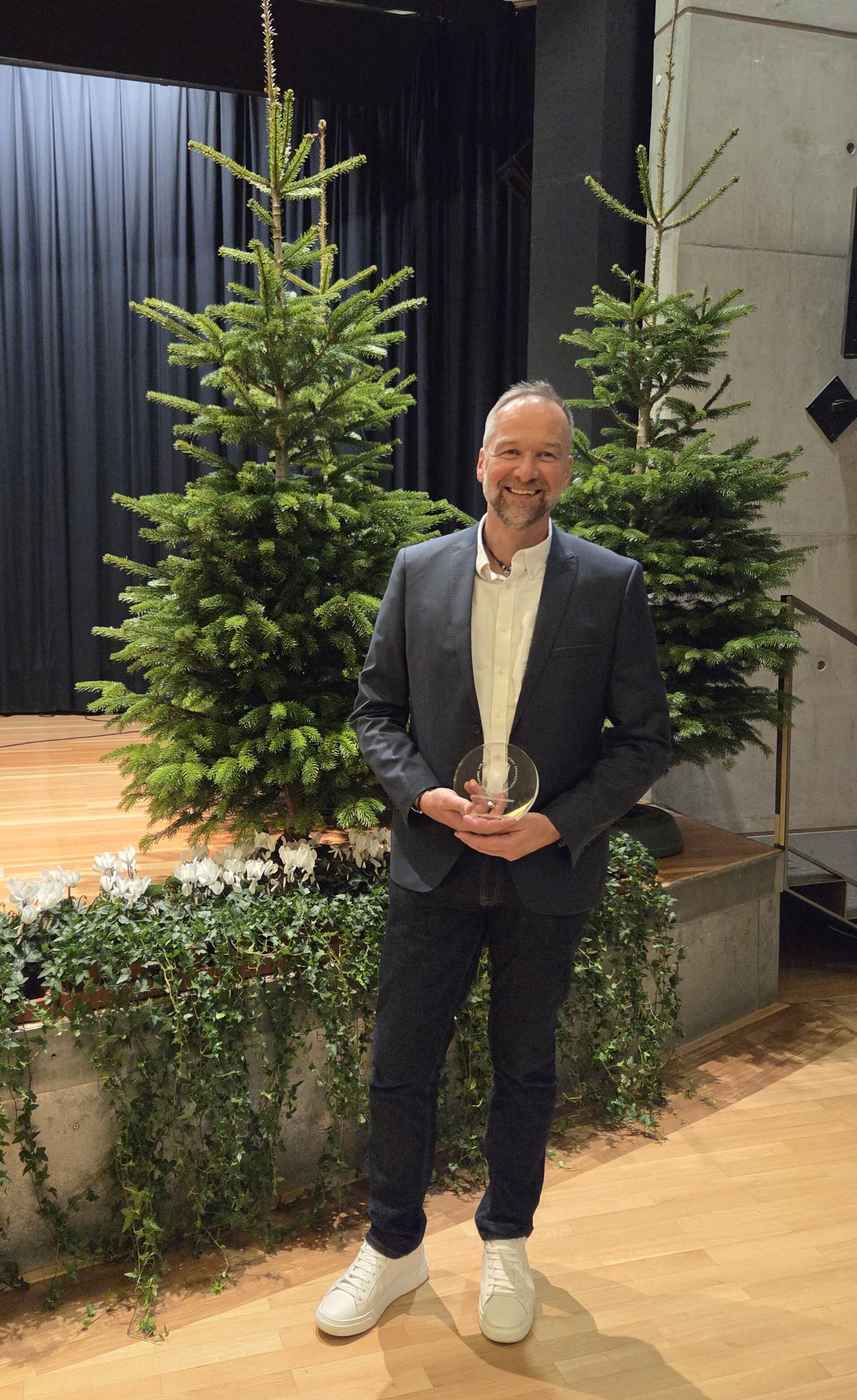 Mann in Anzug h&auml;lt einen Preis und steht vor zwei Weihnachtsb&auml;umen auf einer B&uuml;hnenfl&auml;che.