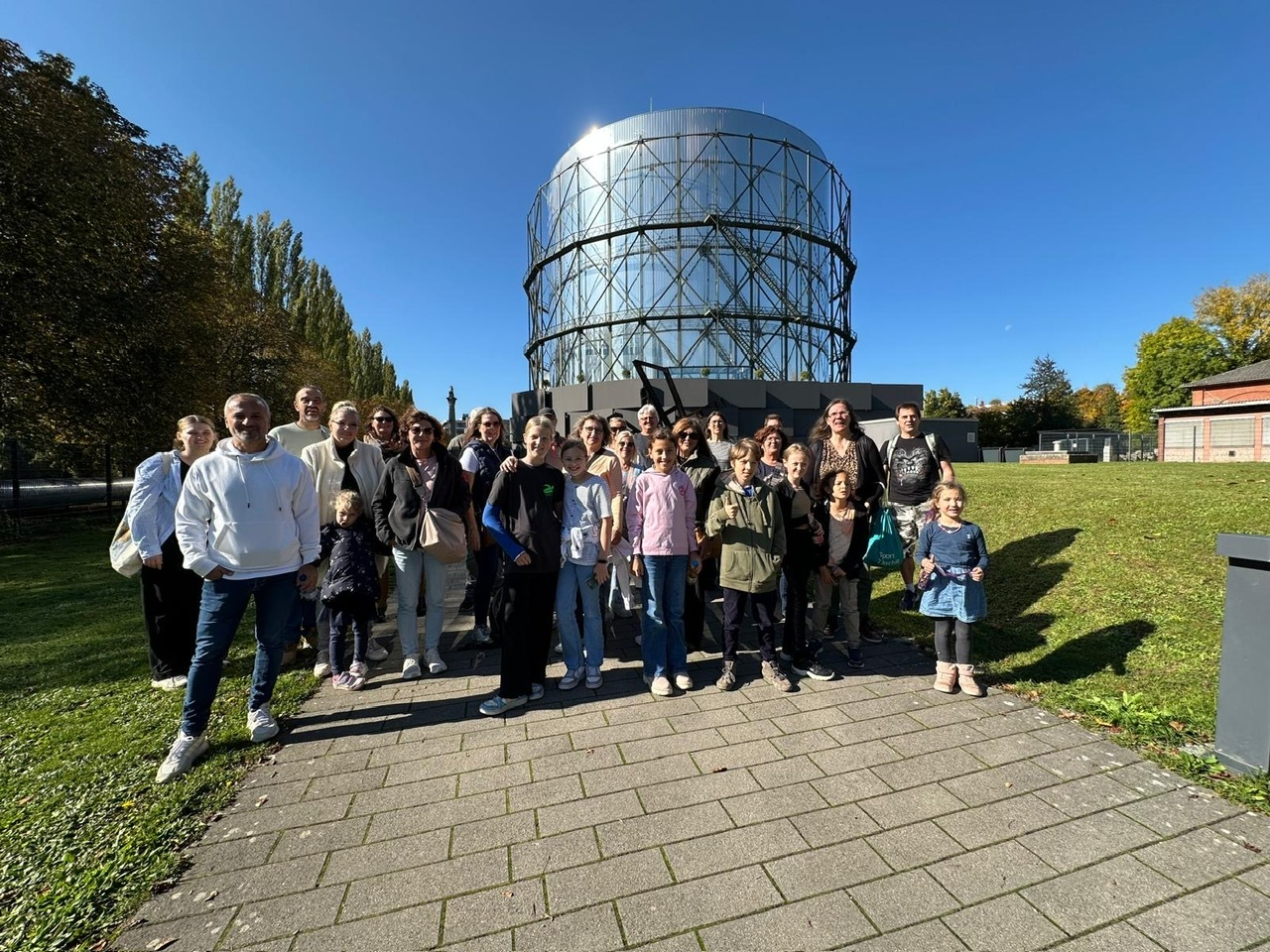 Gruppe von 25 Personen posiert vor einem gro&szlig;en, zylinderf&ouml;rmigen Geb&auml;ude an einem sonnigen Tag.