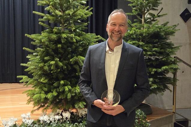 Mann in Anzug h&auml;lt einen Preis und steht vor zwei Weihnachtsb&auml;umen auf einer B&uuml;hnenfl&auml;che.