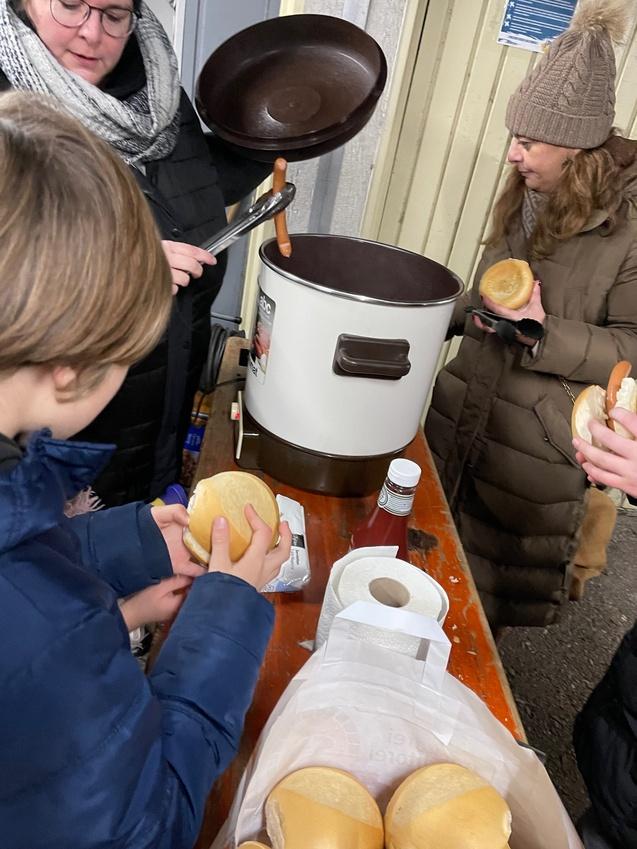 Personen servieren hei&szlig;e W&uuml;rstchen aus einem Topf, w&auml;hrend andere Br&ouml;tchen halten und sich um die Verteilung bem&uuml;hen.