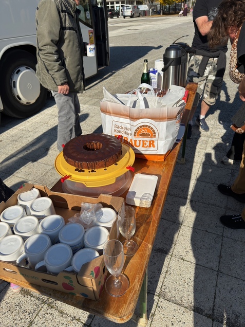 Tisch mit Kuchen, Gl&auml;sern und einer Kaffeekanne; im Hintergrund Menschen und ein Fahrzeug sichtbar.