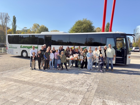 Gruppe von Erwachsenen und Kindern posiert vor einem Reisebus auf einem Platz mit B&auml;umen und blauem Himmel.