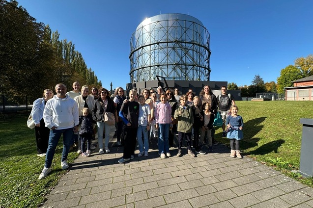 Gruppe von 25 Personen posiert vor einem gro&szlig;en, zylinderf&ouml;rmigen Geb&auml;ude an einem sonnigen Tag.