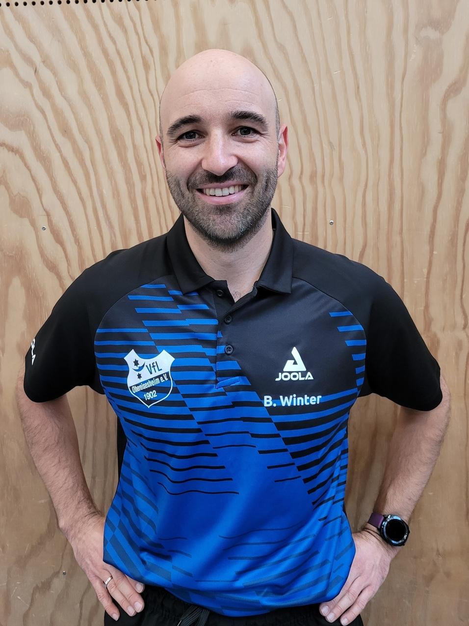 L&auml;chelnder Mann mit Glatze und Polohemd in Blau, steht vor einer holzget&auml;felten Wand und tr&auml;gt eine Armbanduhr.