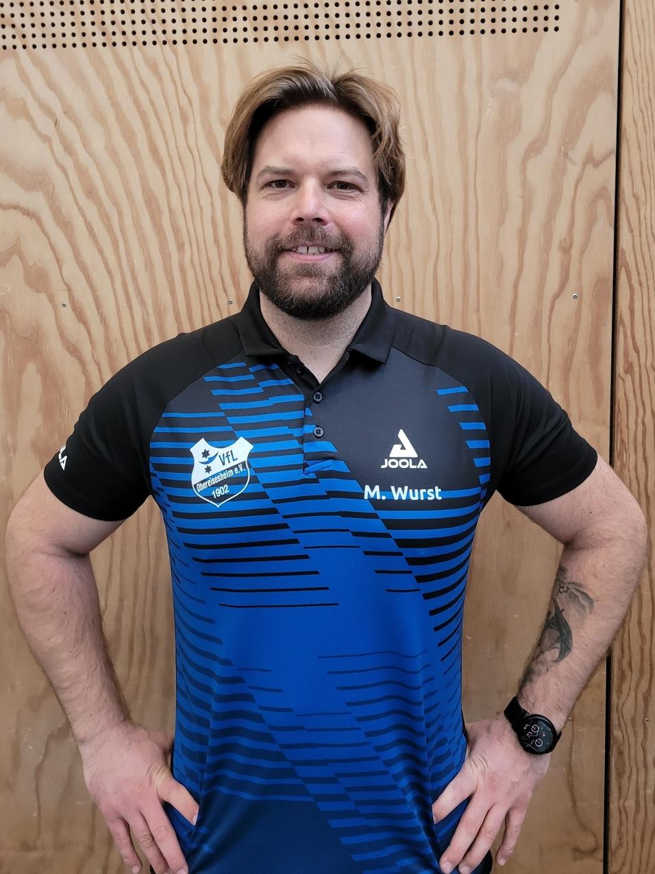 Portr&auml;t eines l&auml;chelnden Mannes in einem blau-schwarzen Poloshirt mit Aufdruck, vor einer holzverkleideten Wand.