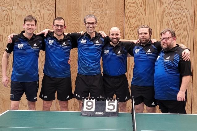 Sechsergruppe von Tischtennisspielern in blauer Sportkleidung vor einem Tisch mit Punktestand 9-5.