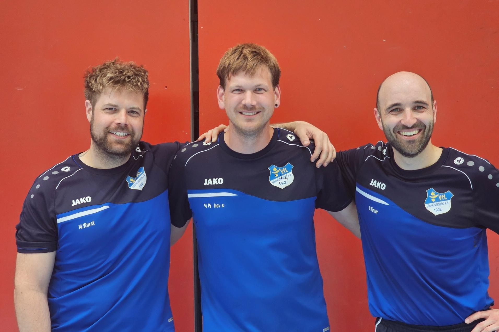 Drei l&auml;chelnde M&auml;nner in blauen Sportshirts posieren freundlich vor einer roten Wand.