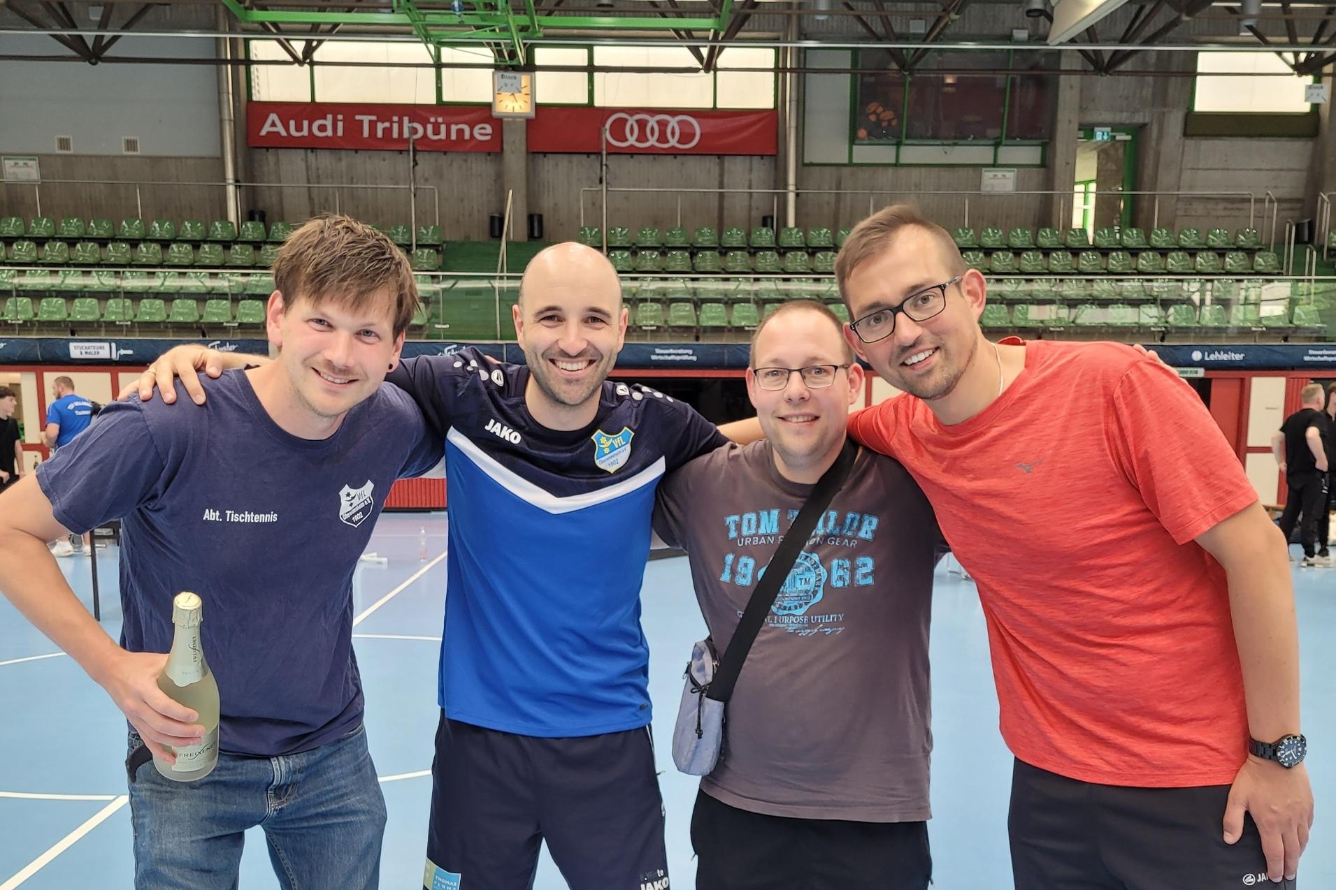 Vier M&auml;nner stehen l&auml;chelnd zusammen in einer Sporthalle, eine Flasche steht im Vordergrund auf dem Boden.