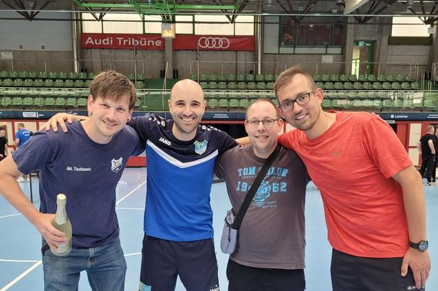 Vier M&auml;nner stehen l&auml;chelnd zusammen in einer Sporthalle, eine Flasche steht im Vordergrund auf dem Boden.