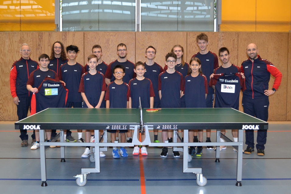 Gruppenfoto einer Tischtennismannschaft mit 15 Spielern und Trainern, stehend vor einer Tischtennisplatte in einer Sporthalle.