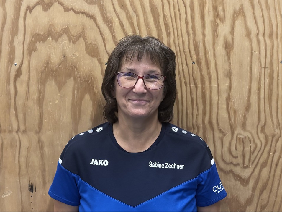 Frau mit Brille und kurzen Haaren tr&auml;gt ein blaues Sporttrikot vor einer Holzdecke.
