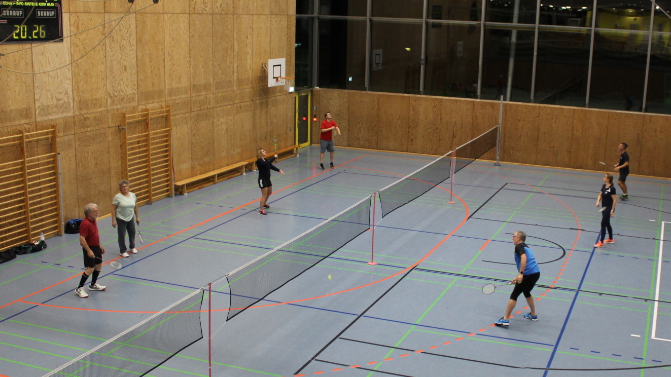 Zwei Gruppen von Spielern trainieren Badminton in einer modernen Sporthalle mit Holzvert&auml;felung.