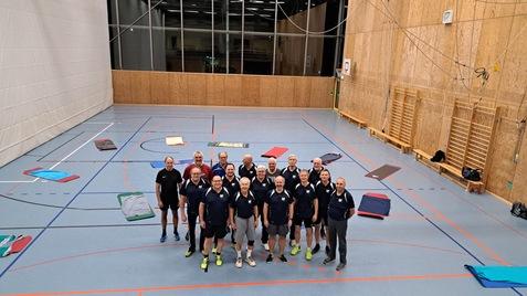 Grupppenfoto von 15 M&auml;nnern in Sportkleidung in einer Turnhalle mit Matten auf dem Boden.
