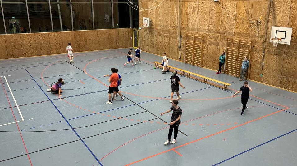 Sporthalle mit mehreren Jugendlichen, die unterschiedliche Spiele auf einem blauen Sportboden spielen.