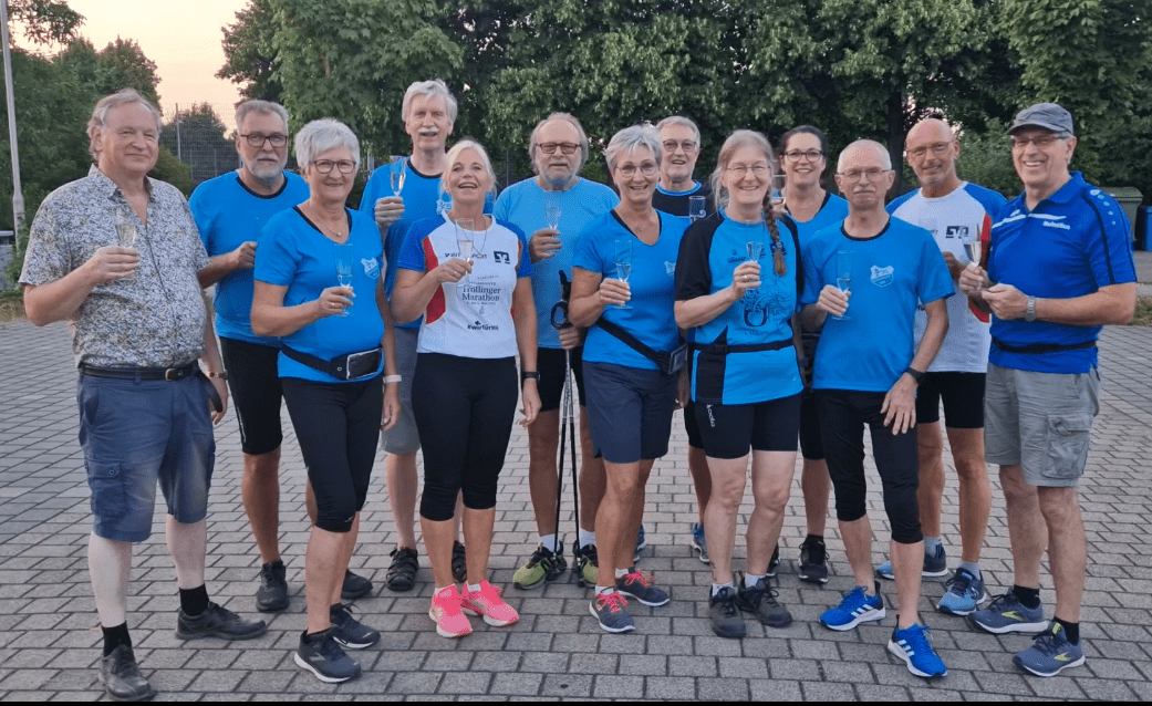 Gruppe von 13 Personen in Sportkleidung, die auf einem Platz stehen und Gl&auml;ser mit Getr&auml;nken halten.