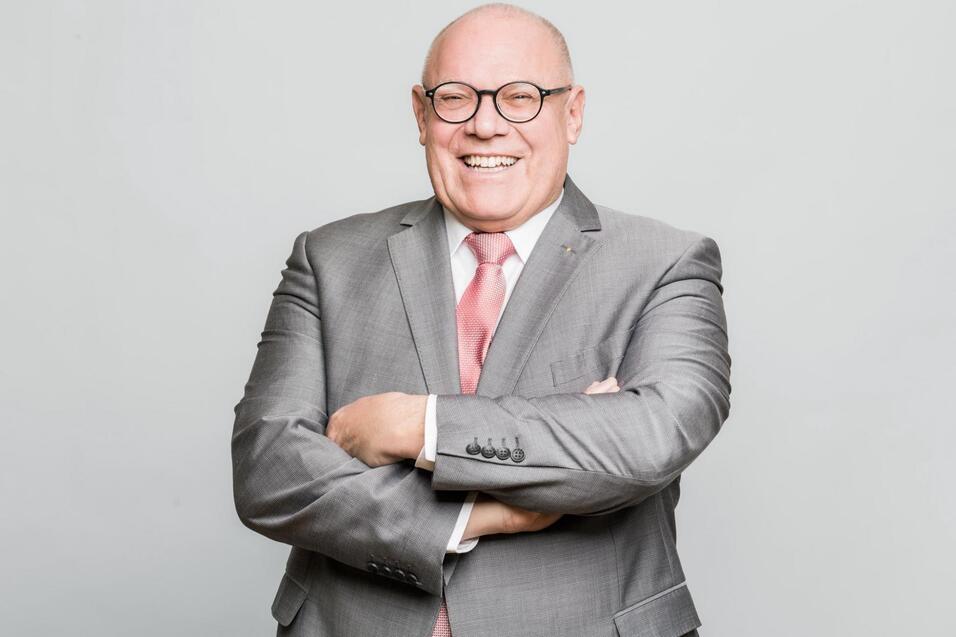 L&auml;chelnder &auml;lterer Mann in Anzug mit Krawatte steht mit verschr&auml;nkten Armen vor einem grauen Hintergrund.