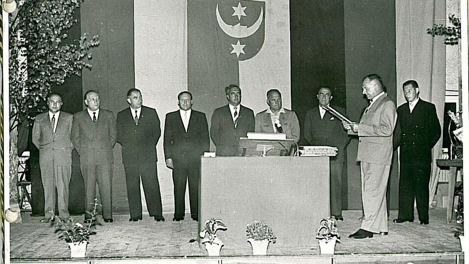 Gruppenfoto von sieben M&auml;nnern bei einer Vereinsgr&uuml;ndung, ein Mann spricht am Podium vor einer Flagge.