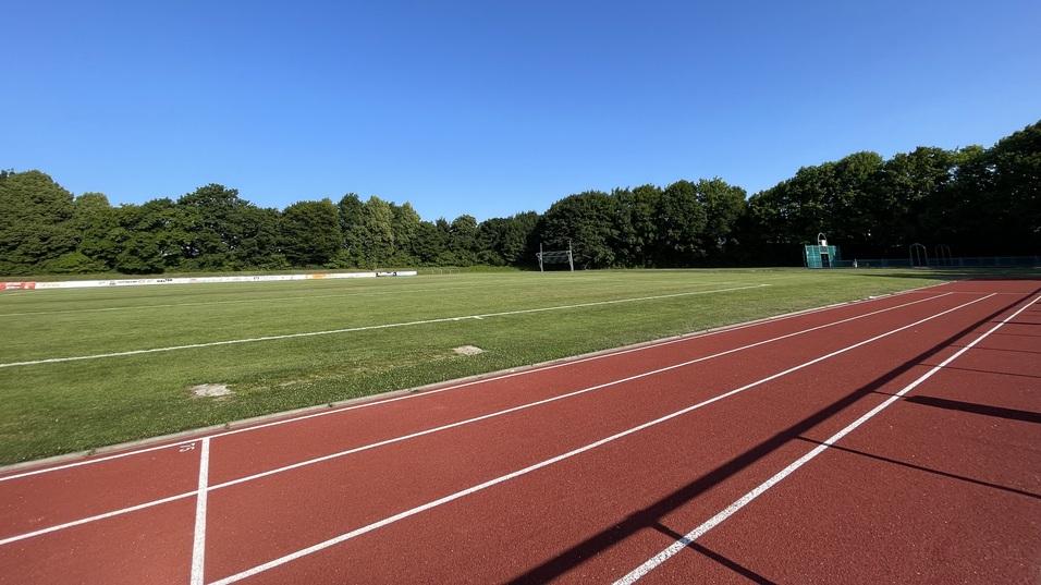 Leere Laufbahn mit rotem Belag und gepflegtem Rasenplatz, umgeben von B&auml;umen unter klarem, blauem Himmel.