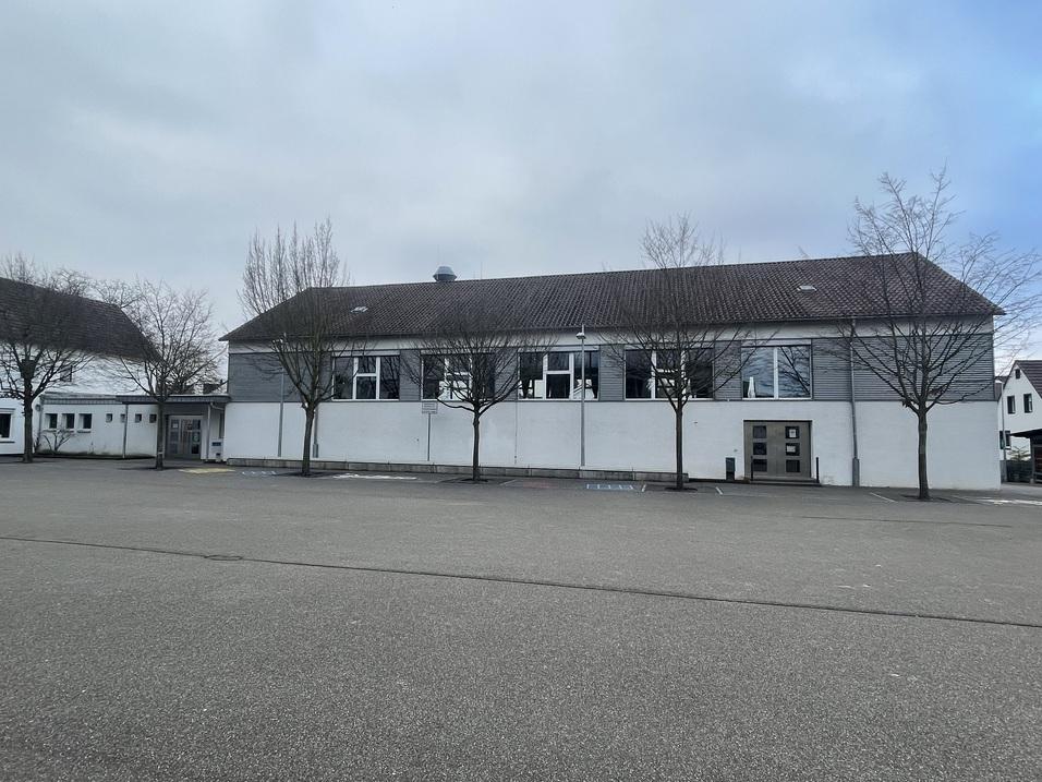 Einfamilienhaus mit grauer Fassade und Fenstern, umgeben von kahlen B&auml;umen auf einem Parkplatz bei bew&ouml;lktem Himmel.