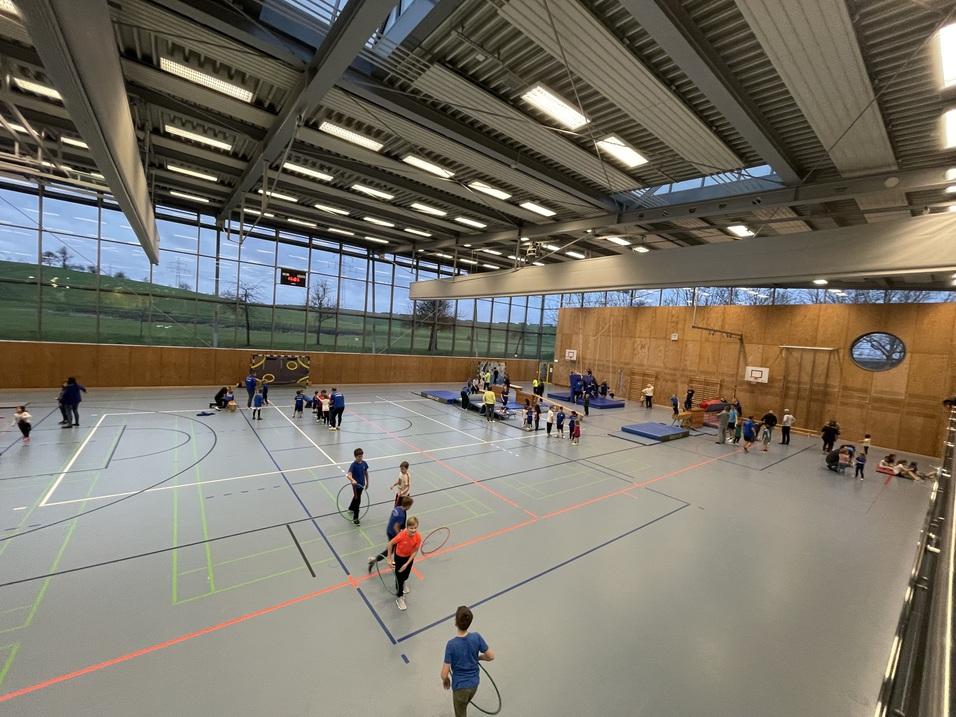Blick in eine Sporthalle mit Kindern, die verschiedene Aktivit&auml;ten ausf&uuml;hren, z.B. mit B&auml;llen und Turnger&auml;ten.