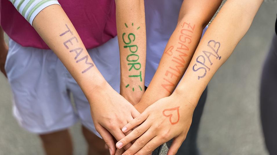 Vier Arme liegen übereinander, mit farbiger Schrift: „TEAM“, „SPORT“, „ZUSAMMEN“, „SPASS“ auf der Haut.