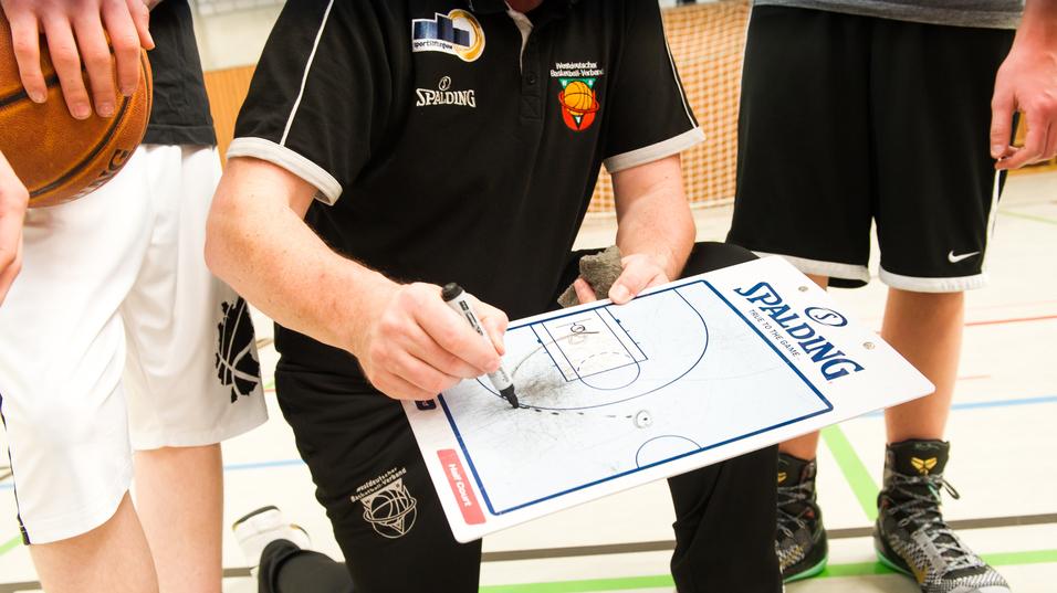 Trainer kniet mit Taktiktafel und zeichnet Spielzüge, während Spieler im Hintergrund stehen und einen Basketball halten.